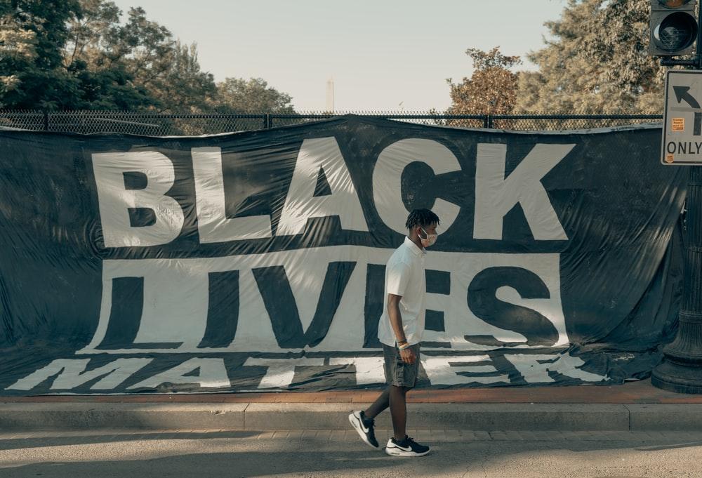 A Man Wearing a Facemask Walking by a Large “Black Lives Matter” Banner