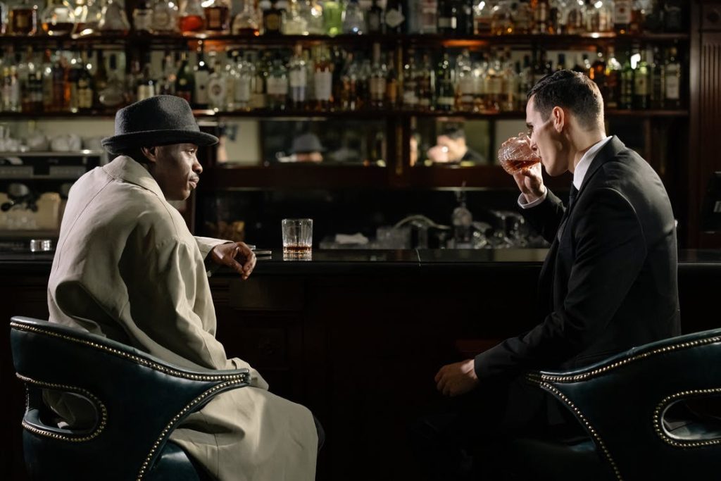 A Caucasian Male in a Suit Sitting at a Bar with an African American Male in a Trench Coat and Hat While Drinking from a Glass