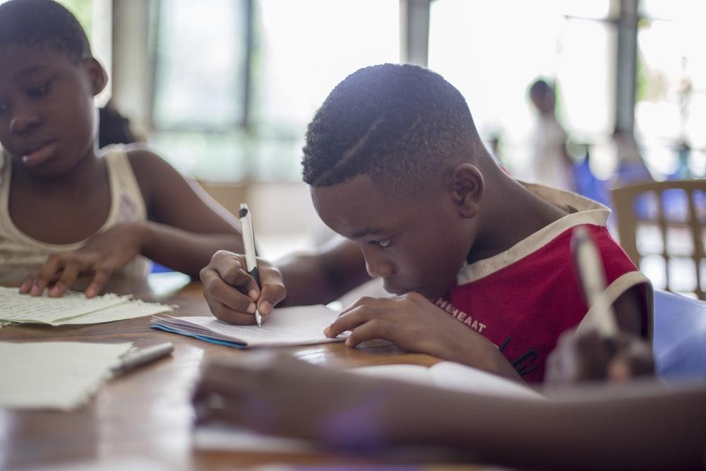 A Little Boy Writing on a Notebook While Sitting at a School Desk